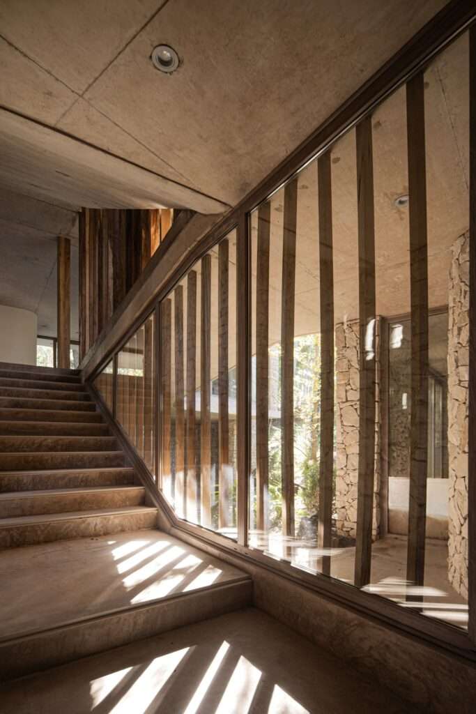 Modern interior concrete staircase with wooden steps, viewed through a glass partition framed by vertical timber posts casting linear shadows.
