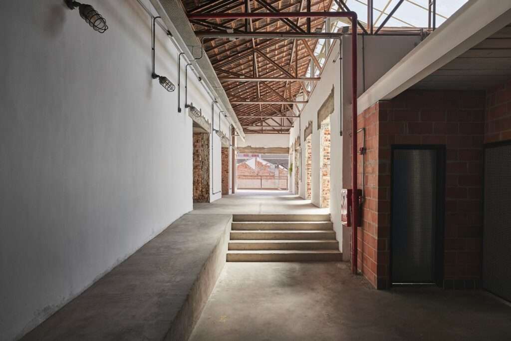 Interior of a transition space within the Fábrica Bhering adaptive reuse in Rio de Janeiro, Brazil. View looking along a ramped hallway towards a landing with steps and large windows. The space features whitewashed walls, exposed industrial lighting, and a red brick wall section. Photographed by Rafael Salim.