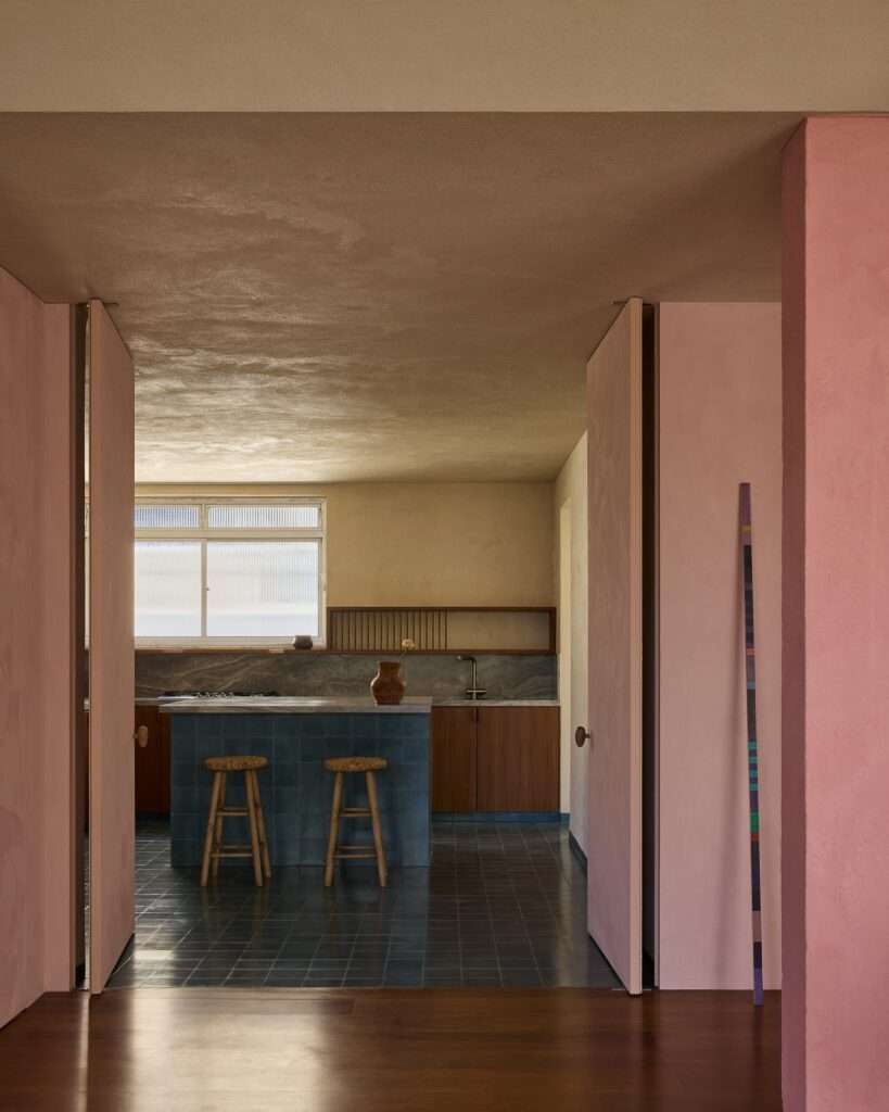 View through a pink-plastered opening into an open kitchen with a blue tiled island, wooden bar stools, dark tiled floor, and wood cabinetry under a frosted glass window.