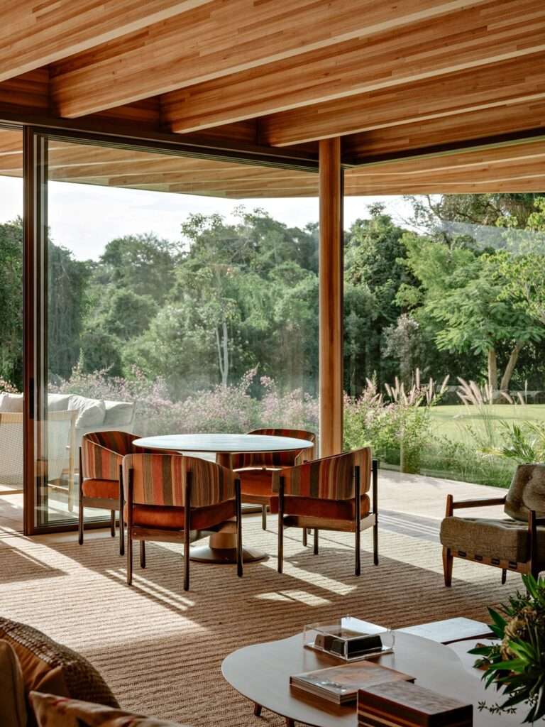 Interior view of a smaller lounge area in AMS House featuring a round wooden table, striped upholstery chairs, and large glass windows framing the outdoor gardens.