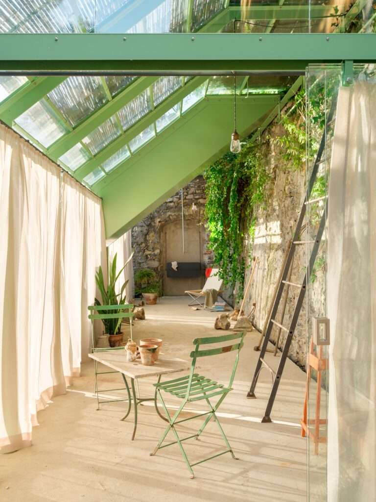 An interior photograph looking down the length of the greenhouse during daytime, showing a bright, transparent environment with glass walls and roof panels framed in light green metal. Sunlight filters through semi-transparent curtains drawn along the glass facade. At the far end, a green bistro table and matching metal chairs sit on the concrete floor near an old wooden ladder against the stone wall.