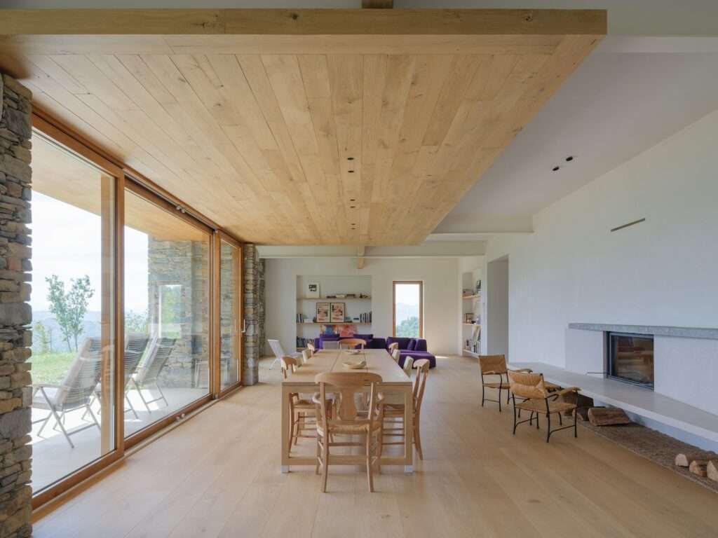 Bright minimalist dining room featuring a long solid wood table, large sliding glass doors opening to the landscape, and an overhead oak wood ceiling panel.