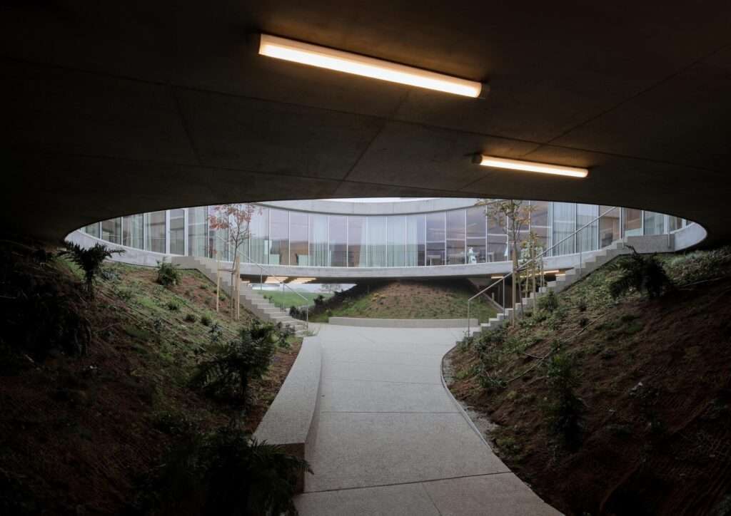 Covered outdoor walkway beneath a low concrete ceiling leading toward the brightly lit central circular patio surrounded by planted embankments.