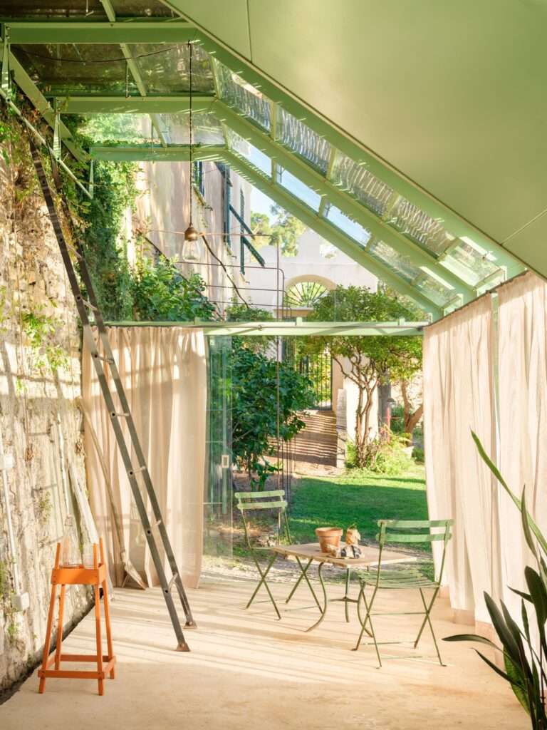 An interior perspective of the greenhouse taken during daytime, looking down its length from the far end toward the garden entrance. Light green metal framing and glass panels create a sun-drenched corridor lined with light curtains. An old wooden ladder rests against the historical stone wall on the left, while a green bistro table and chairs sit on the concrete floor in the center, framing the view to the outdoor garden beyond.