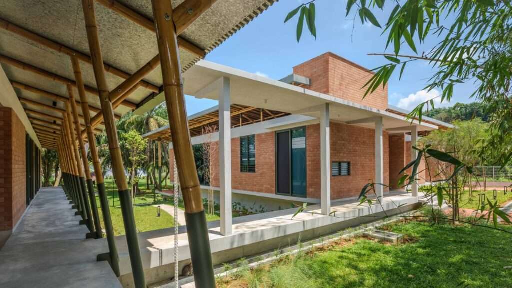 Perspective view of Anjung House showing connected concrete walkways, terracotta brick volumes, a bamboo roof canopy, and a landscaped green garden under a blue sky.