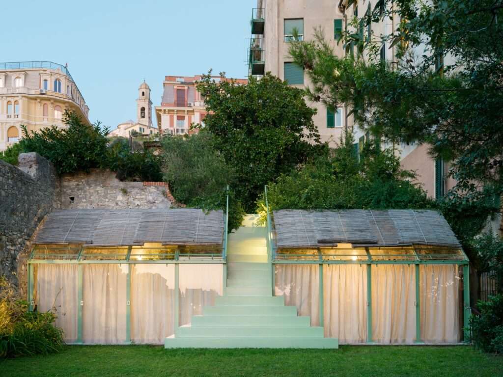 A full front elevation photograph of the greenhouse taken during dusk. The two glass-roofed modules with drawn curtains are illuminated from within by warm indoor lighting, framing the central green staircase. The historic stone wall behind and the surrounding lush vegetation are set against a clear twilight sky with historic apartment towers in the background.