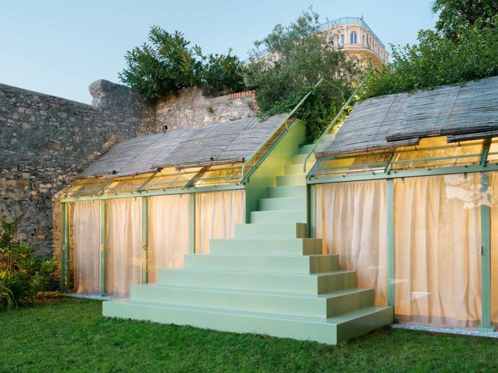 An angled perspective photograph of the greenhouse taken at dusk, focusing on the central green painted staircase and the illuminated glass structure to the right. Warm interior light glows through the curtains and glass panels, contrasting with the ancient stone wall behind it and the soft evening light on the lawn in the foreground.