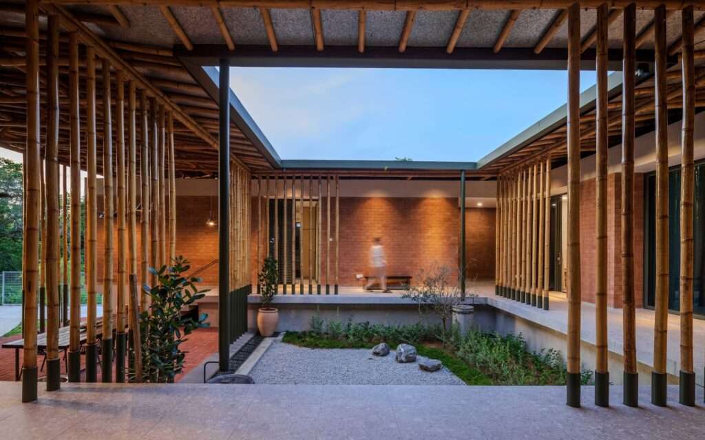 Central courtyard of Anjung House at twilight, featuring an open-to-sky gravel garden surrounded by brick walls, bamboo screens, and warm interior lighting.
