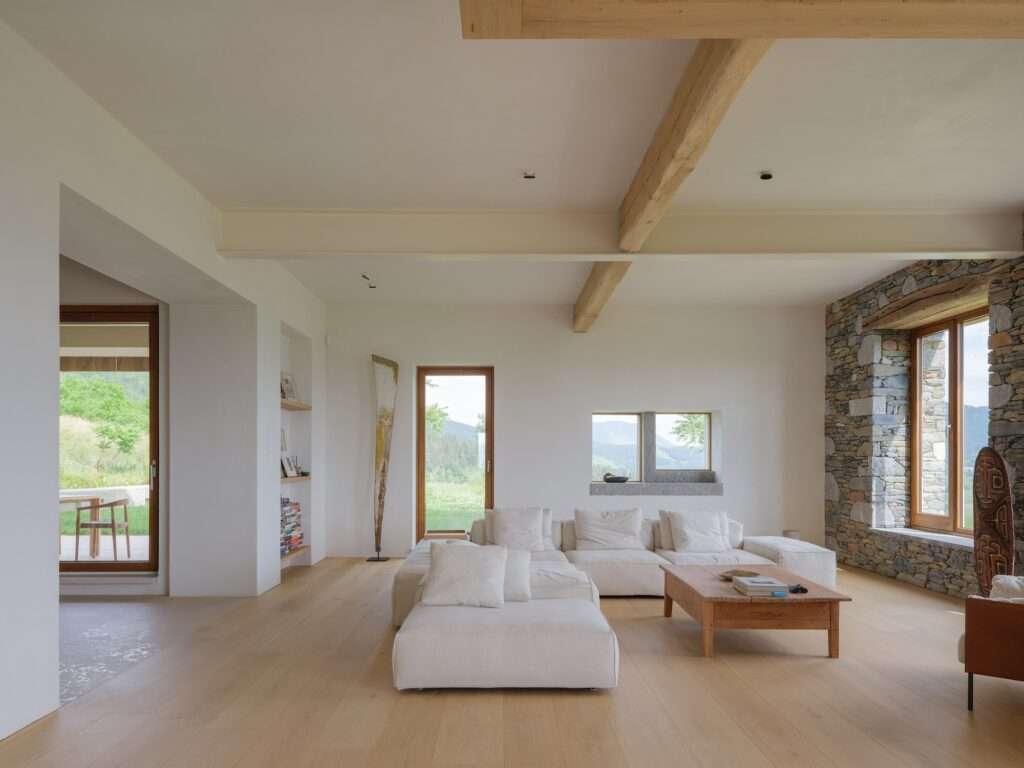 Contemporary living room with a white sectional sofa, solid wood coffee table, exposed structural timber columns, and deep stone window sills overlooking rolling hills.