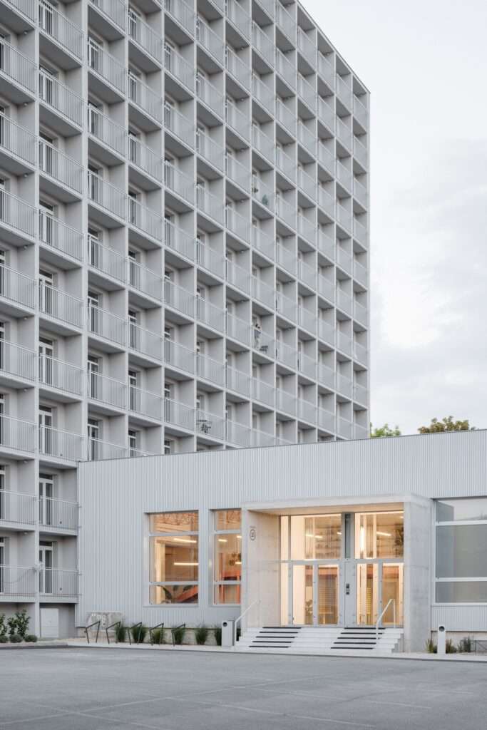 Dusk view of the concrete entrance canopy and glass doors of Terasa housing complex under rows of illuminated modular balconies.