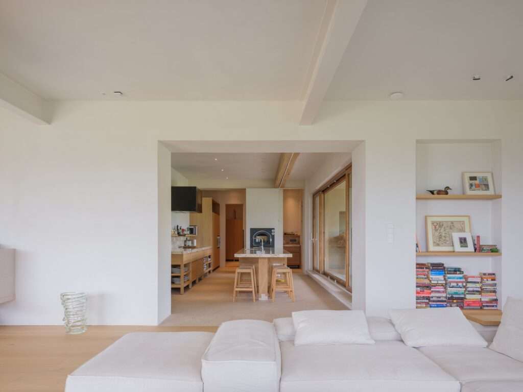 View from a white living space looking into a minimalist modern kitchen with wood cabinetry, an island, and stools framed by a thick structural wall opening.