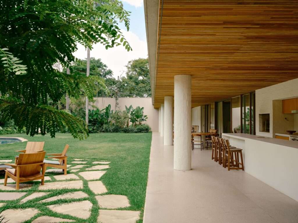 Outdoor patio bar and kitchen area of Jacarezinho II House with wooden barstools, circular brick pillars, and stone stepping pathways across a lawn.