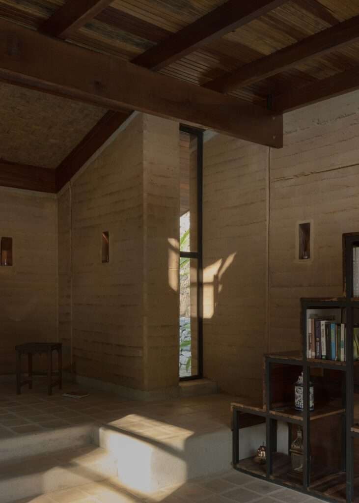 A narrow, vertical window in a rammed earth wall creates a sharp, geometric pattern of sunlight and shadow across the floor and walls.