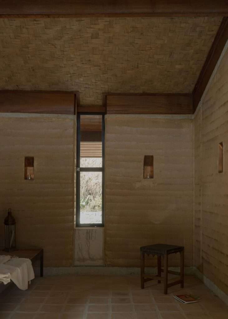 An interior corner view showing a single bed, a small wooden stool, a lantern, and a narrow vertical window in a rammed earth wall.