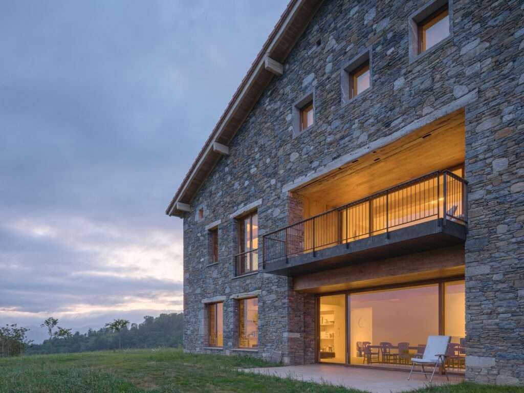 Evening twilight view of the Casa Basque House exterior stone wall with warm interior lights glowing through large glass windows and an integrated upper balcony.