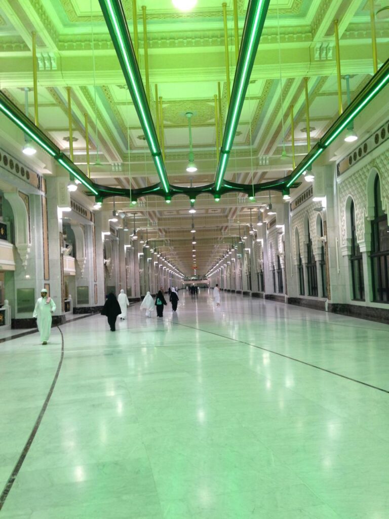 Interior view of the expanded corridors in Al-Masjid Al-Haram, featuring polished marble floors, arched columns, and modern green linear ceiling lighting.