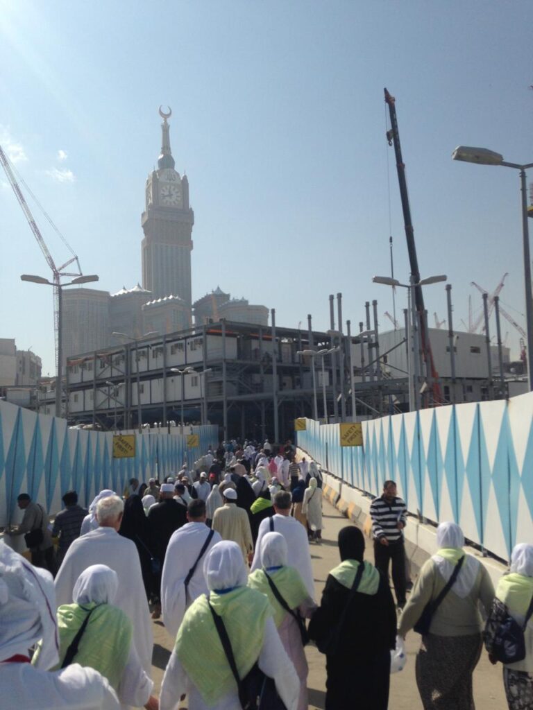Crowds of pilgrims in white Ihram walking along a temporary fenced pedestrian bridge during the construction phase of Al-Masjid Al-Haram, with the Makkah Clock Tower in the background.