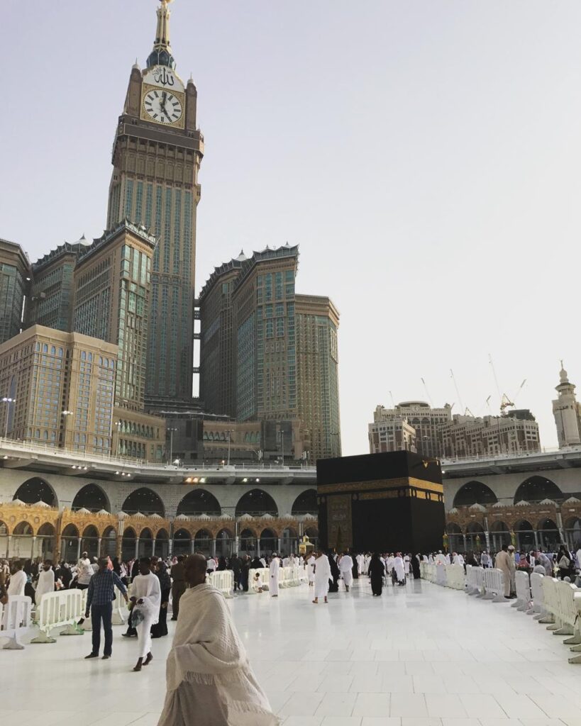 The holy Kaaba surrounded by pilgrims performing Tawaf during the day, enclosed by a temporary elevated walkway and the towering Abraj Al Bait clock tower in the background.