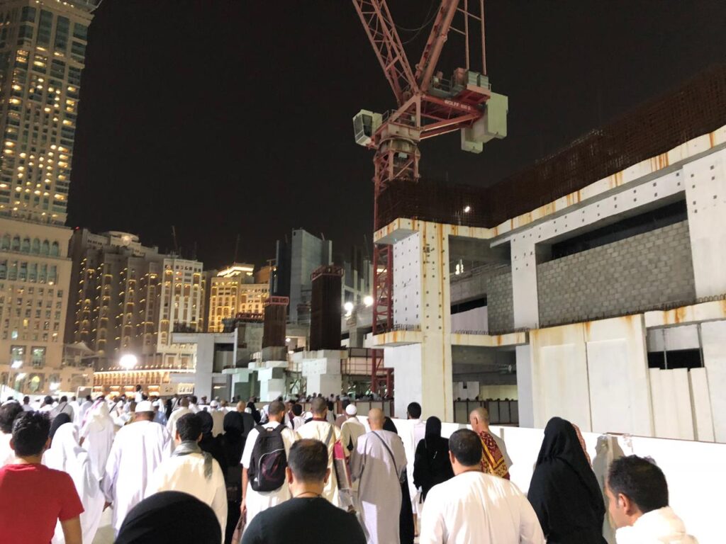 Thousands of pilgrims gathered around the Kaaba at night, with multiple massive construction cranes operating in the background over partially built mosque structures.