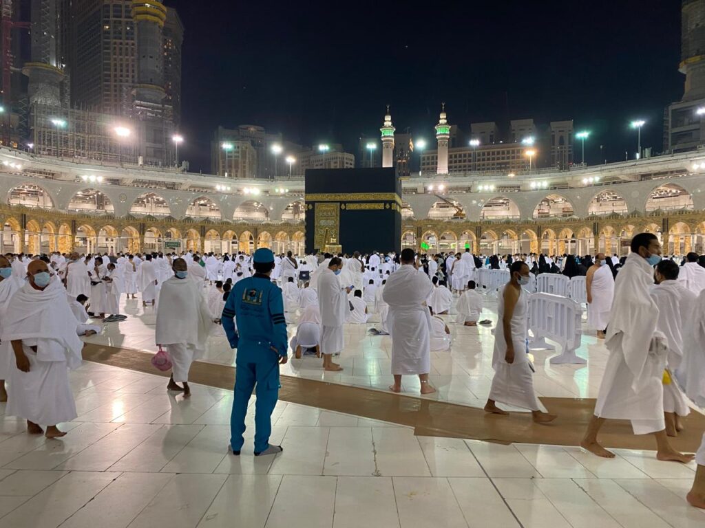 Pilgrims walking at night past a massive construction zone with exposed concrete pillars, steel rebar, and a towering red crane inside the Grand Mosque complex.
