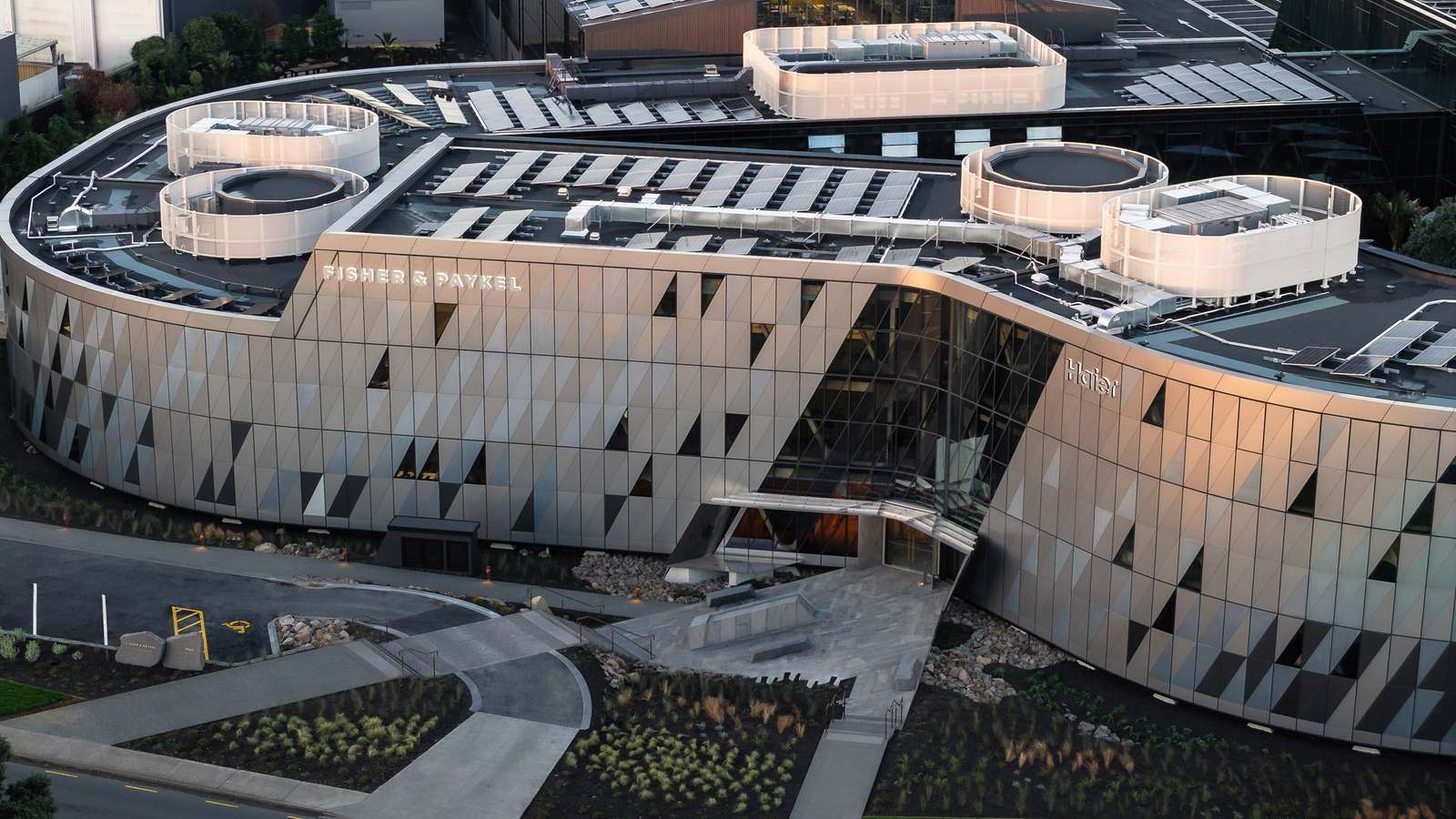 Aerial view of a three-story curved office building with rooftop solar panels and branding.
