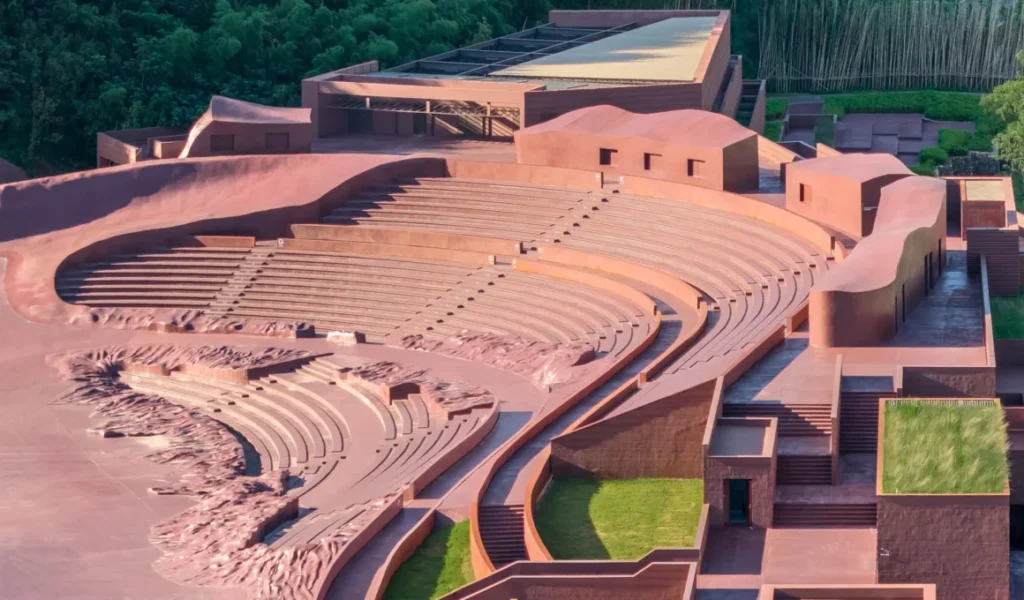 Aerial view of a red concrete sunken amphitheater with curved seating steps and adjacent geometric buildings.