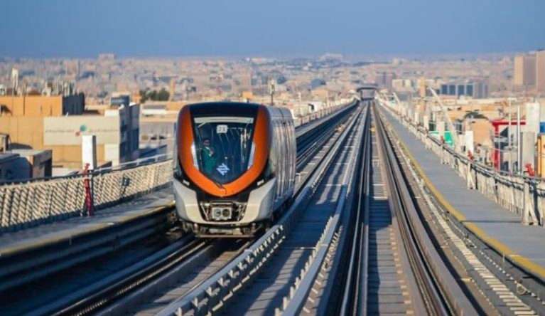 An orange and grey Riyadh Metro train moving along an elevated track with urban cityscape in background.