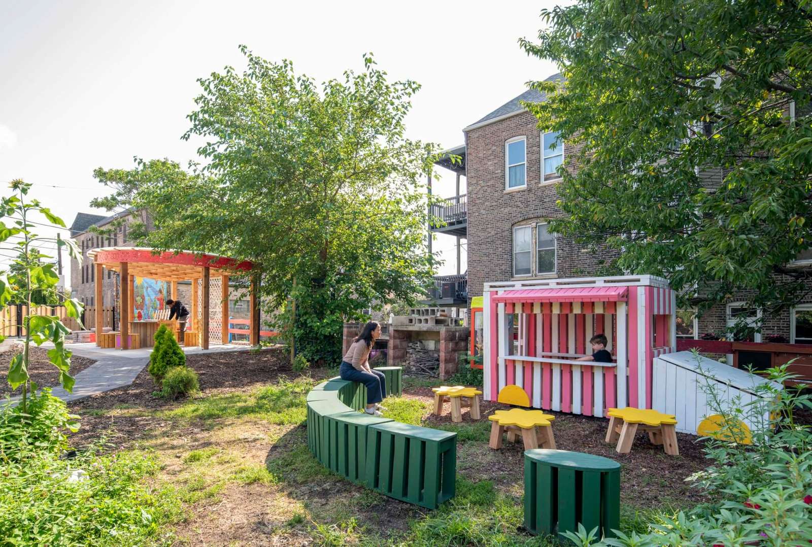 Outdoor community garden featuring timber play structures, curved green seating bench, and shaded pergola structure.