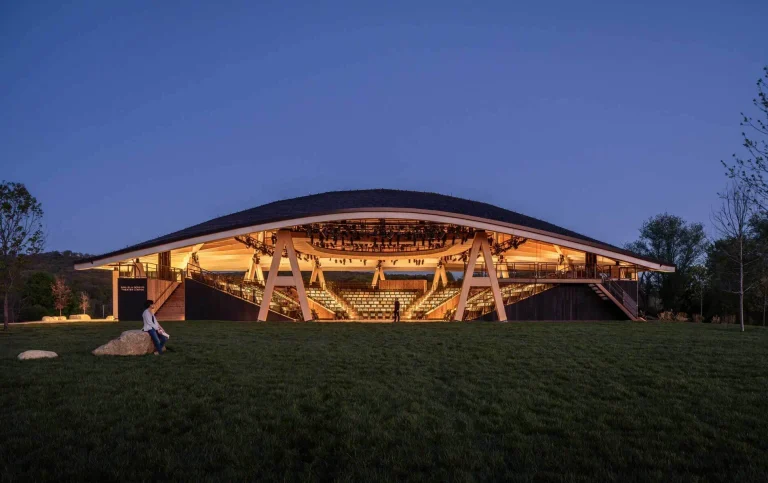 Illuminated open-air mass timber theater bowl and canopy structure set against grassy terrain at dusk.