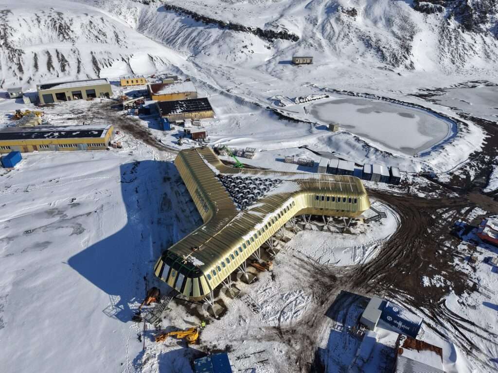 Aerial perspective of the raised golden research building set against snow-draped Antarctic mountains.