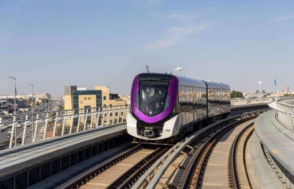 قطار باللونين البنفسجي والفضي يعبر مسارًا مرتفعًا منحنيًا بجوار طريق رئيسي في الرياض.