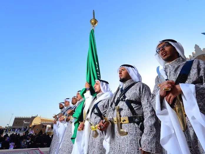 مؤدون بالزي التقليدي يحملون العلم الأخضر أمام واجهة مبنى طيني تقليدي.