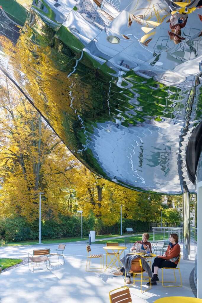 Pedestrians seated outdoors beneath a mirrored undulating roof overhang reflecting surrounding nature