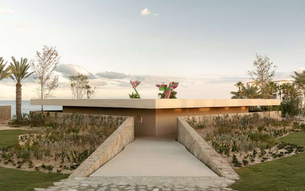 Straight-on exterior view of a low concrete volume with an entrance ramp flanked by stone walls.