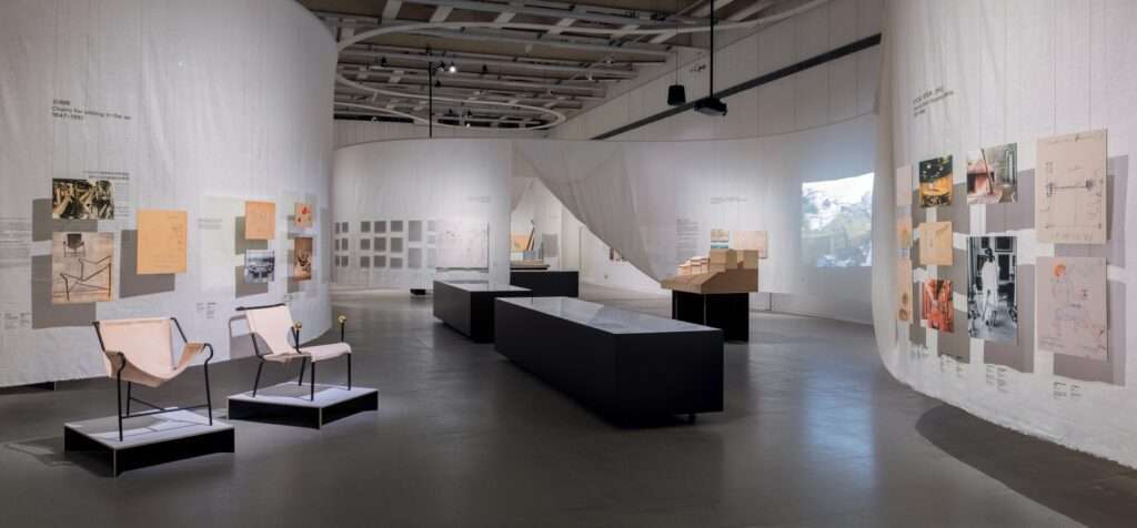 Continuous white linen curtains forming curved exhibition spaces with displayed furniture and sketches.