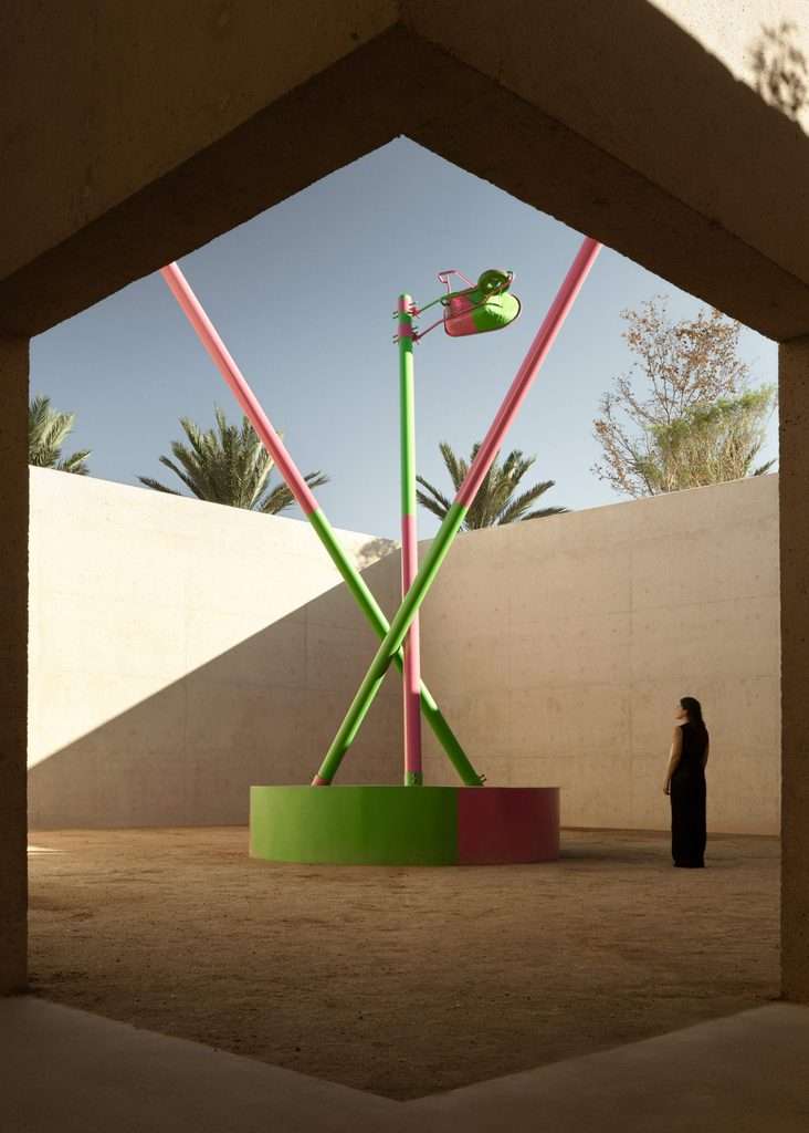 View framed by concrete structure showing a sand-covered courtyard containing a colorful tall sculpture and a person.