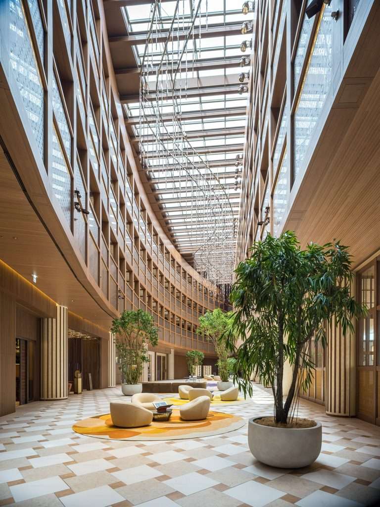 Multi-storey internal atrium with glass skylight ceiling, timber-framed walls, and potted indoor trees.