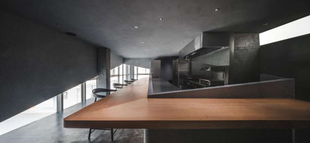 Perspective looking down a long angled wooden dining counter toward an open stainless-steel kitchen.