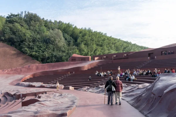 Visitors walking along a reddish concrete pathway inside a large open-air amphitheater with curved seating tiers.
