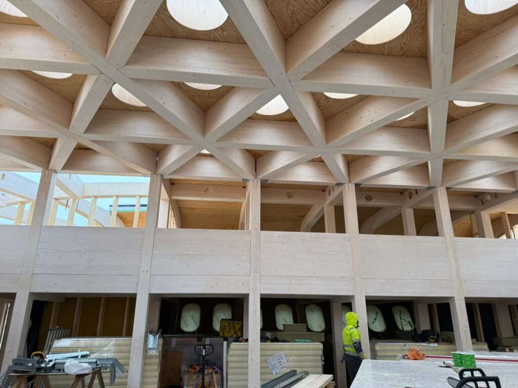 Interior under construction featuring exposed mass-timber columns, beams, and triangular ceiling grid with round skylights.