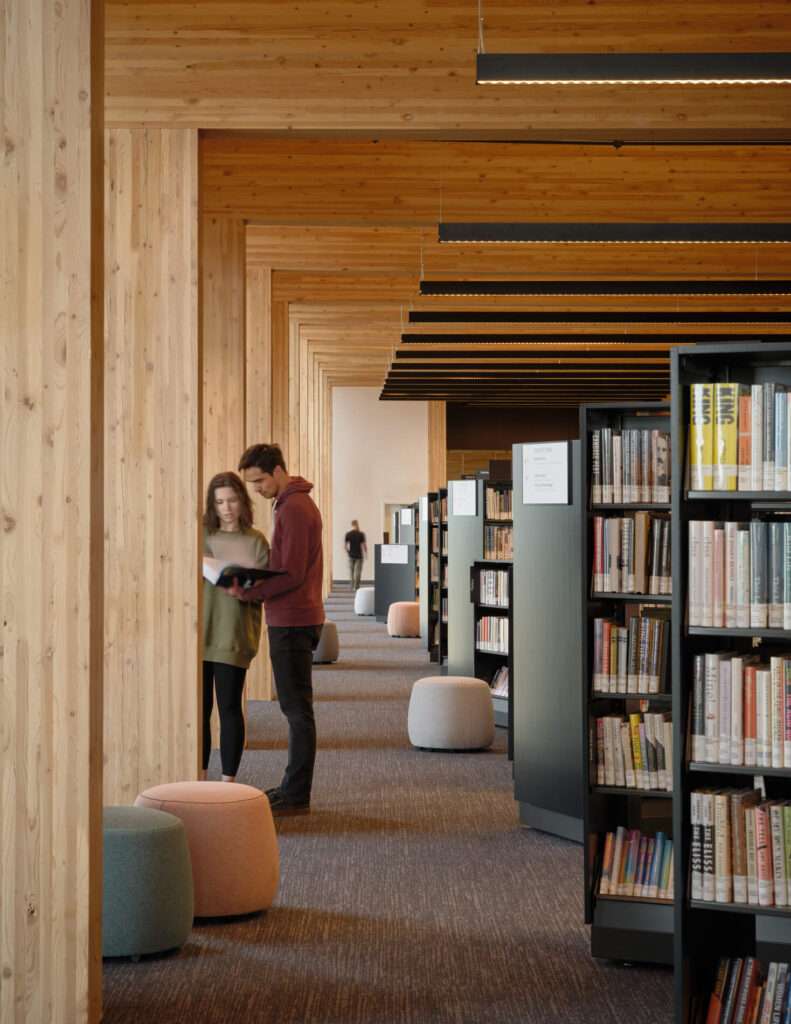 Library aisle flanked by mass timber columns and dark book display shelving.
