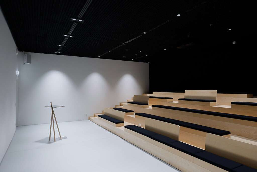 Minimalist auditorium space with tiered wood benches and black padded seating surfaces