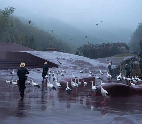 Performers and birds standing on a damp red concrete surface near organic woven structures under a misty sky.