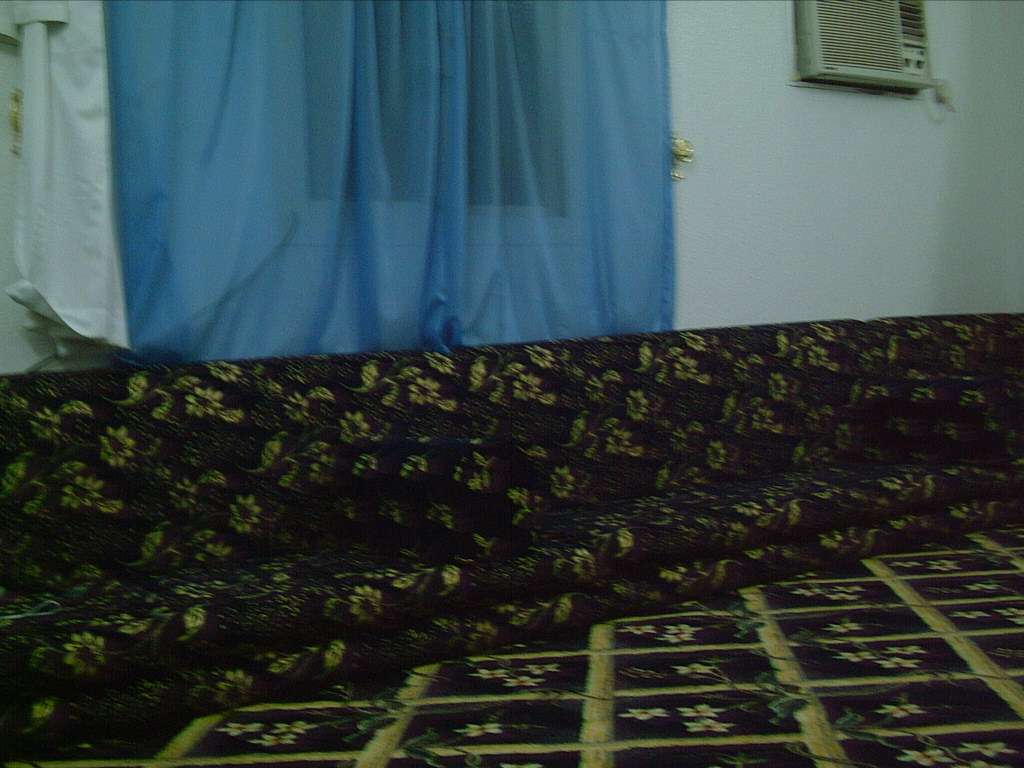 A dimly lit, modest room featuring traditional dark fabric floor seating with a yellow floral pattern, a matching patterned rug, a bright blue window curtain, and an old wall-mounted air conditioning unit.