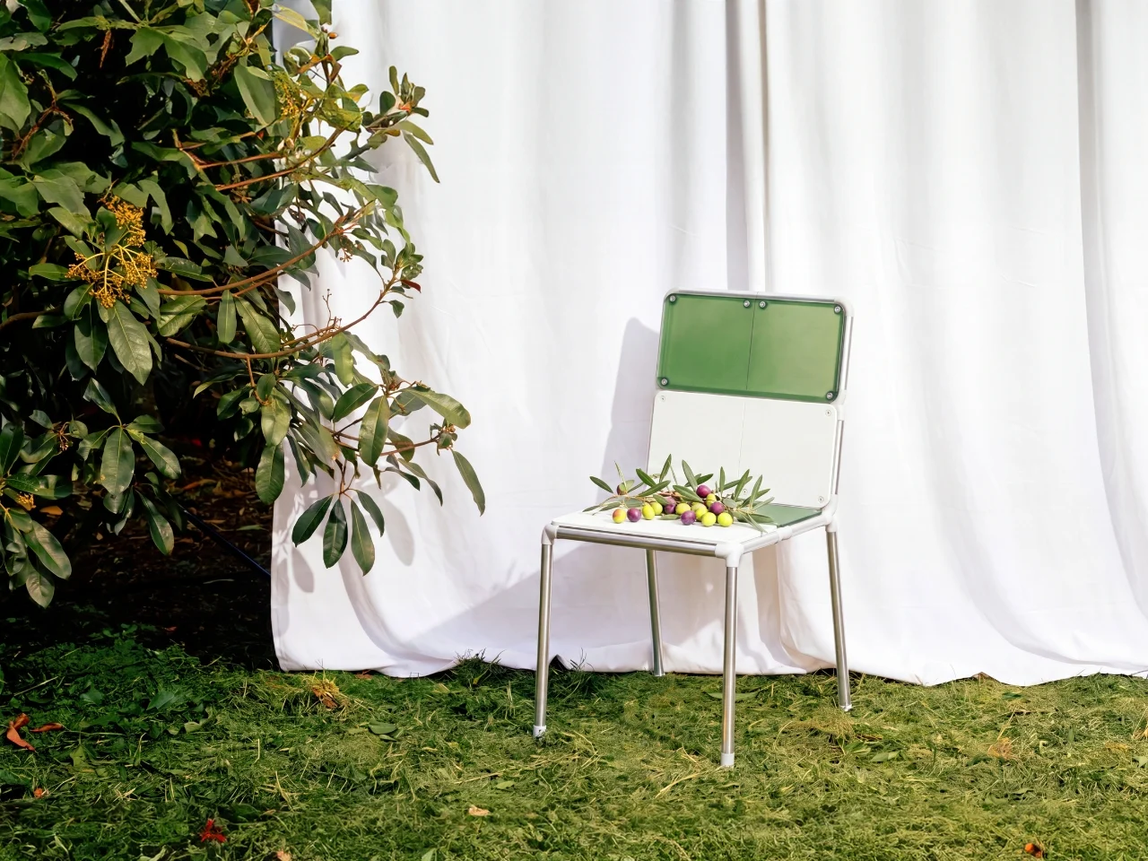 Facade chair with green and white panels, visible fasteners and a tubular frame, placed on grass in front of a white curtain.