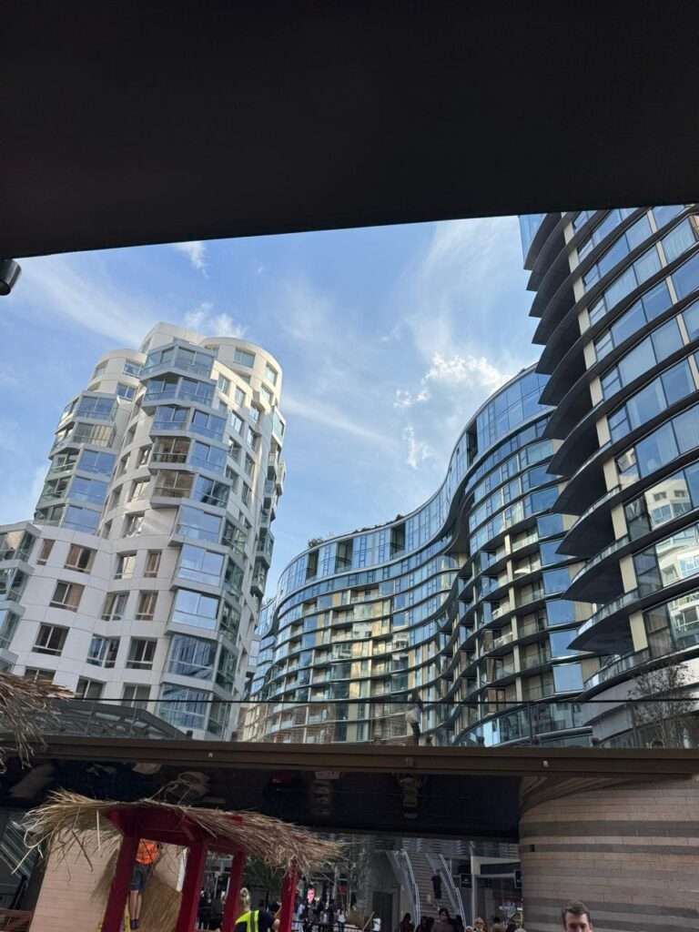 تؤطر هذه الصورة مشروع (Prospect Place) لفرانك جيري، لتعرض واجهته البيضاء المتموجة المميزة بجانب هيكل زجاجي منحني. وفي الخلفية، تلوح إحدى المداخن الشهيرة لمحطة طاقة باترسي في سماء ساطعة، مما يسلط الضوء على تقاطع العمارة النحتية الحديثة والتراث الصناعي.