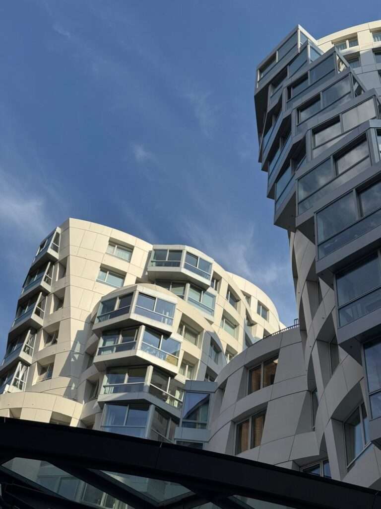 يسلط هذا المنظر المتجه للأعلى الضوء على الحوار المعماري بين واجهتين متناقضتين في المشروع. على اليسار، تنحني الشبكة الهيكلية البيضاء المرنة لجيري إلى الخارج، بينما يتميز الجانب الأيمن بكتلة سكنية أغمق، وأكثر صرامة (ثباتاً)، ومتدرجة بشكل كبير.