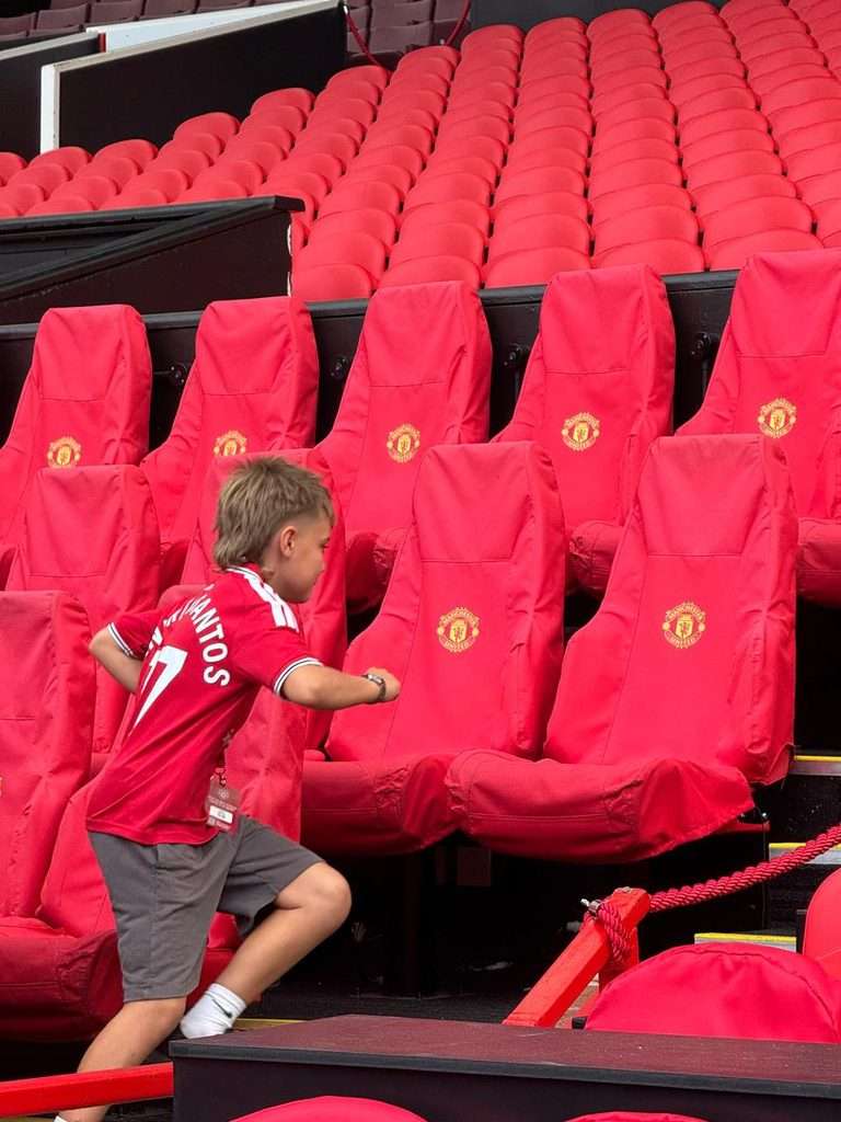 A young boy in a Manchester United football shirt runs towards red padded stadium seats