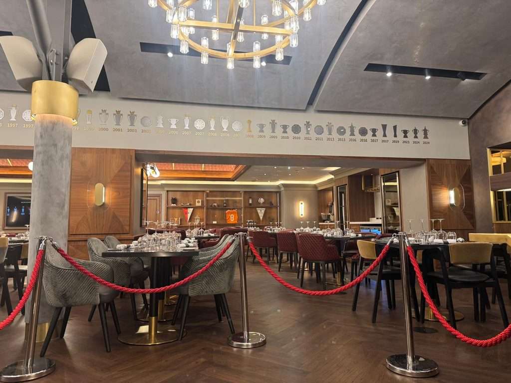 Upscale restaurant dining room with set tables, red stanchion ropes, and a timeline of football trophies along the wall
