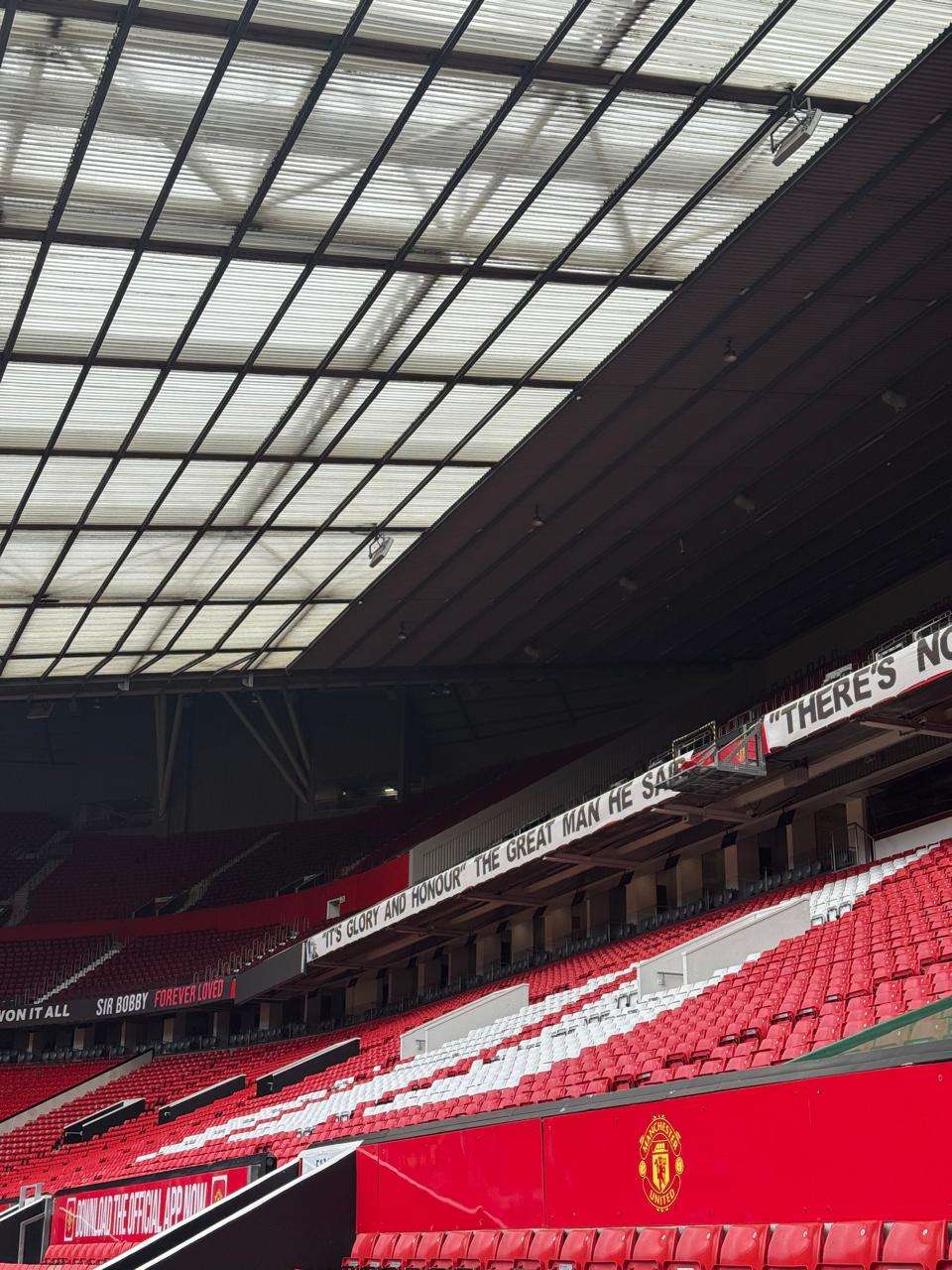 Inside Old Trafford stadium featuring red and white seating, translucent roof, and banners honouring Sir Bobby Charlton